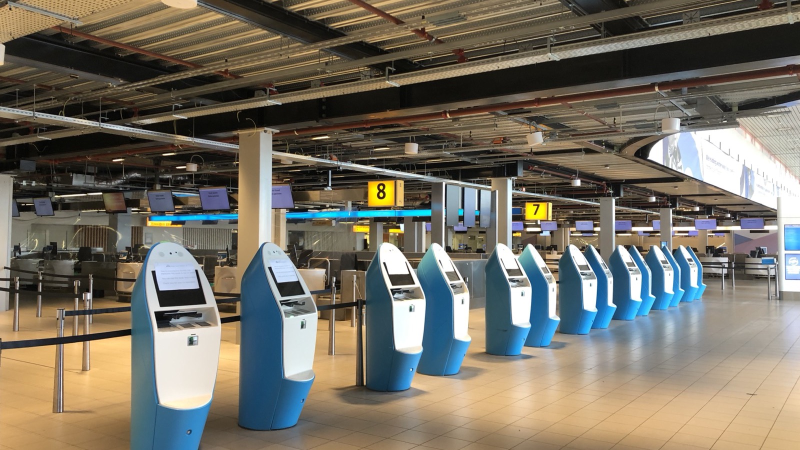 Een lege vertrekhal op Schiphol met een rij blauwe incheckzuilen.